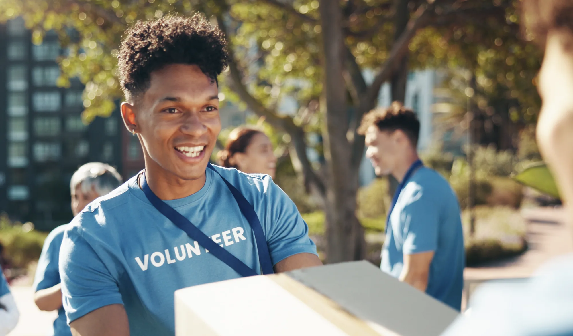 Young man volunteering for Goodwill at an outdoor event, carrying a box and wearing a blue 'Volunteer' shirt.