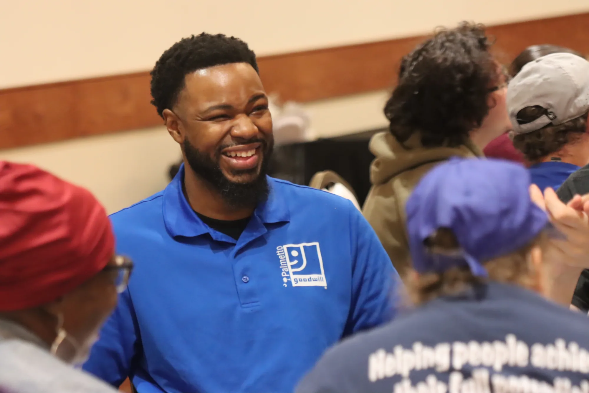 Man in a Palmetto Goodwill uniform smiling while interacting with others during a job training or community program.