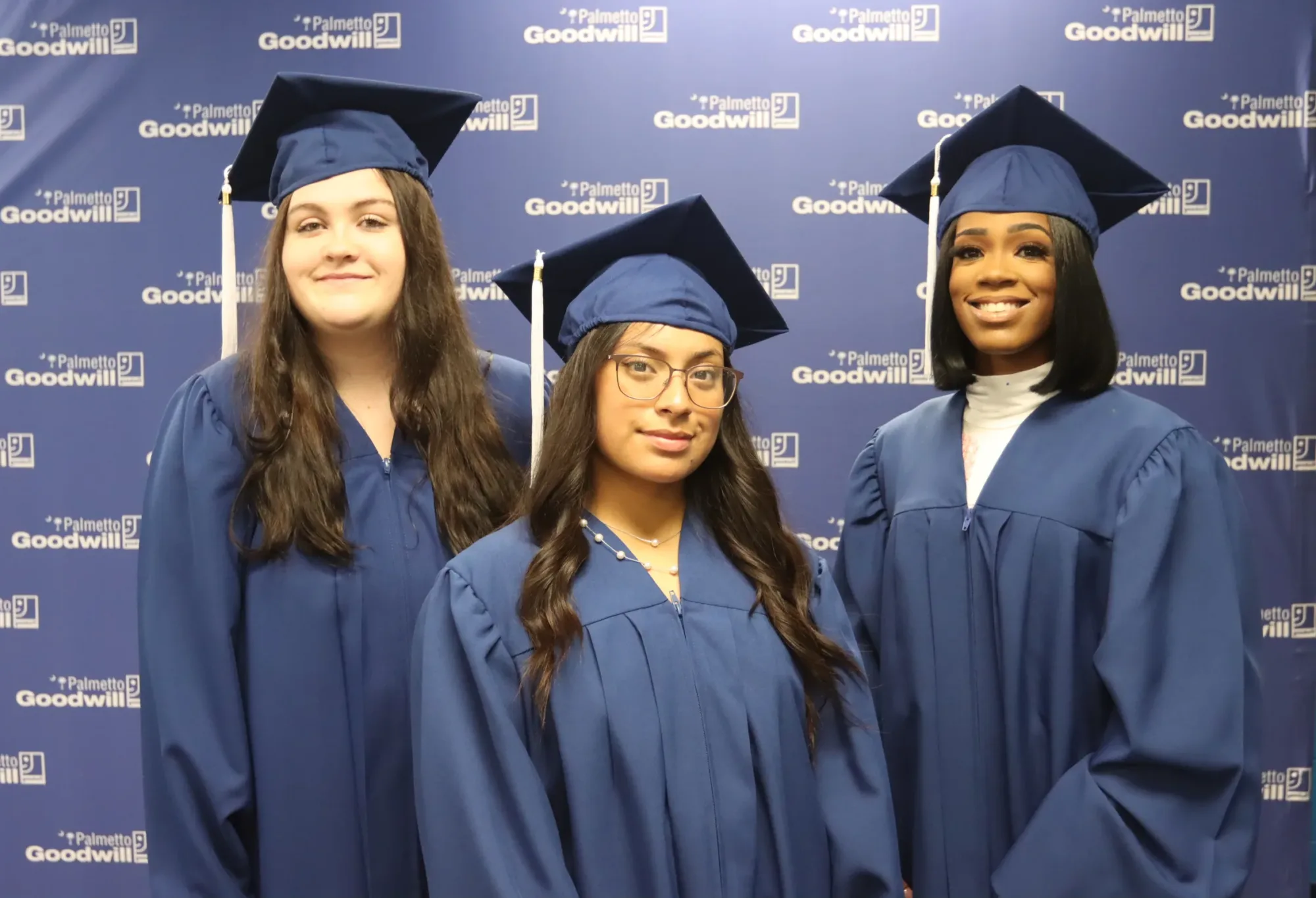 Three diverse female graduates of The Palmetto Excel Center, a tuition-free high school for adults, wearing blue caps and gowns.