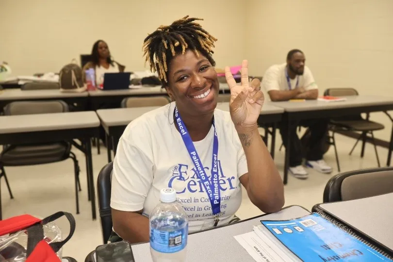 Smiling adult student at The Palmetto Excel Center giving a peace sign while sitting at her desk in class.