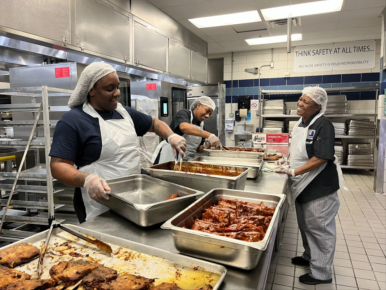 Diverse food service team members, employed through Palmetto Goodwill Contracts, smiling while preparing large trays of food in a professional kitchen.