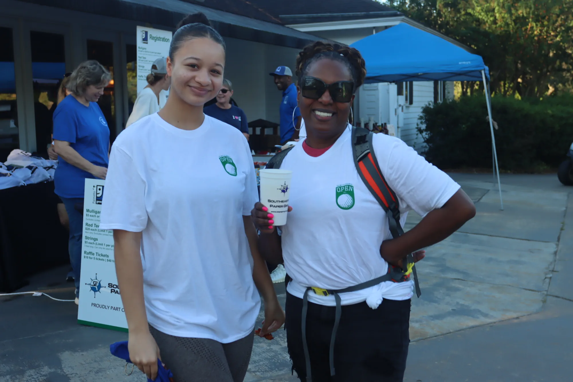 Two women, Palmetto Goodwill volunteers, pose for a photo outside, holding a cup from the event while standing near a registration table and blue tent.