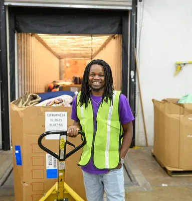 Smiling Palmetto Goodwill production center employee standing with a pallet jack and large box.