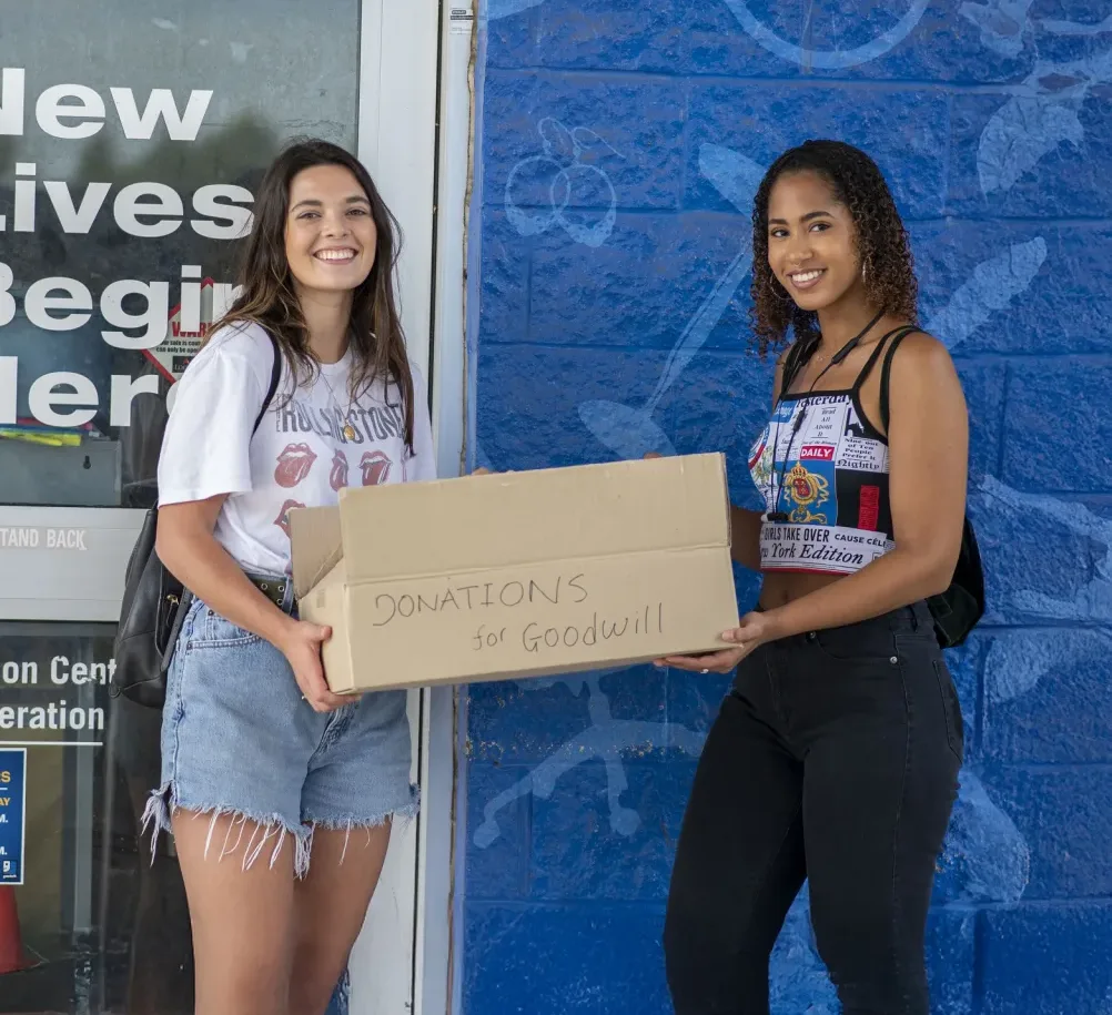 Smiling donors dropping off a box of clothing donations at the Palmetto Goodwill retail store entrance.