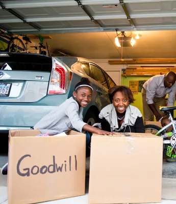 Family in a garage packing two large boxes labeled 'Goodwill' with toys and a bicycle for donation.