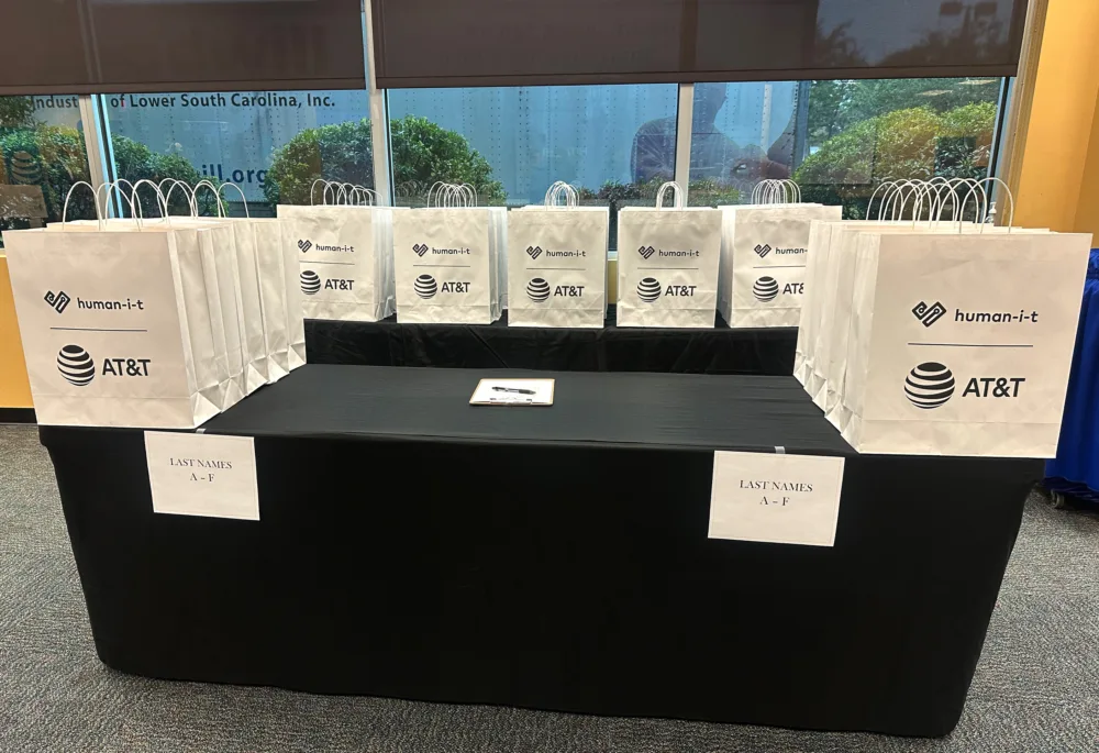 Table set with giveaway bags from AT&T, Human-I-T, and Palmetto Goodwill, showing readiness to distribute laptops for Digital Access Job Training SC.