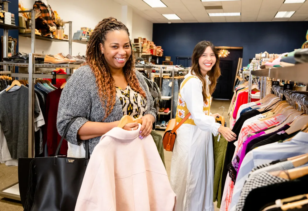 Two women smiling while shopping for high-quality secondhand outerwear and dresses in a well-organized boutique (emphasizing the type of structured garments that benefit from intentional care and storage).