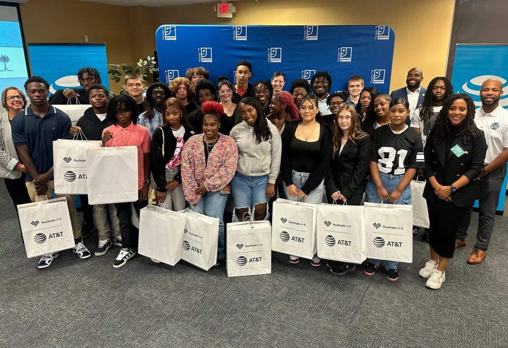 A large group of students holding bags with donated technology in front of a Palmetto Goodwill banner (working toward closing digital divide SC).