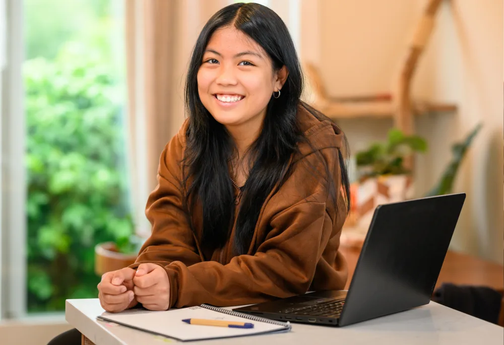 A smiling young student sitting with her refurbished laptop and notebook, representing the success of sustainable technology initiatives in South Carolina.