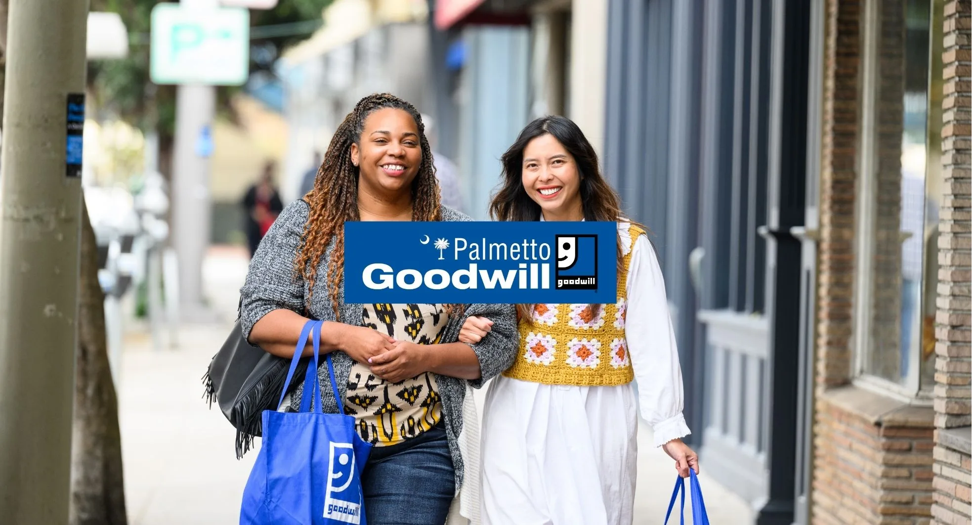 Two smiling women walk down a sunny sidewalk carrying blue Palmetto Goodwill reusable shopping bags (representing a successful, sustainable thrift trip for Earth Month).