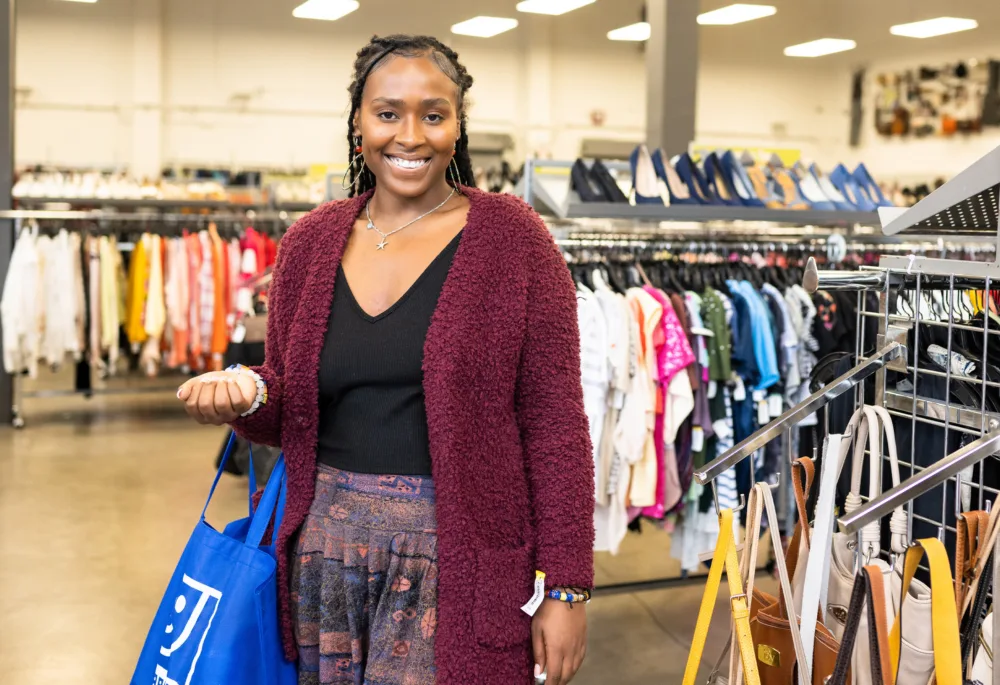 A smiling Black woman with braids, wearing a maroon fuzzy cardigan and unique skirt, stands in a Palmetto Goodwill, presenting beaded bracelets and a reusable shopping bag.