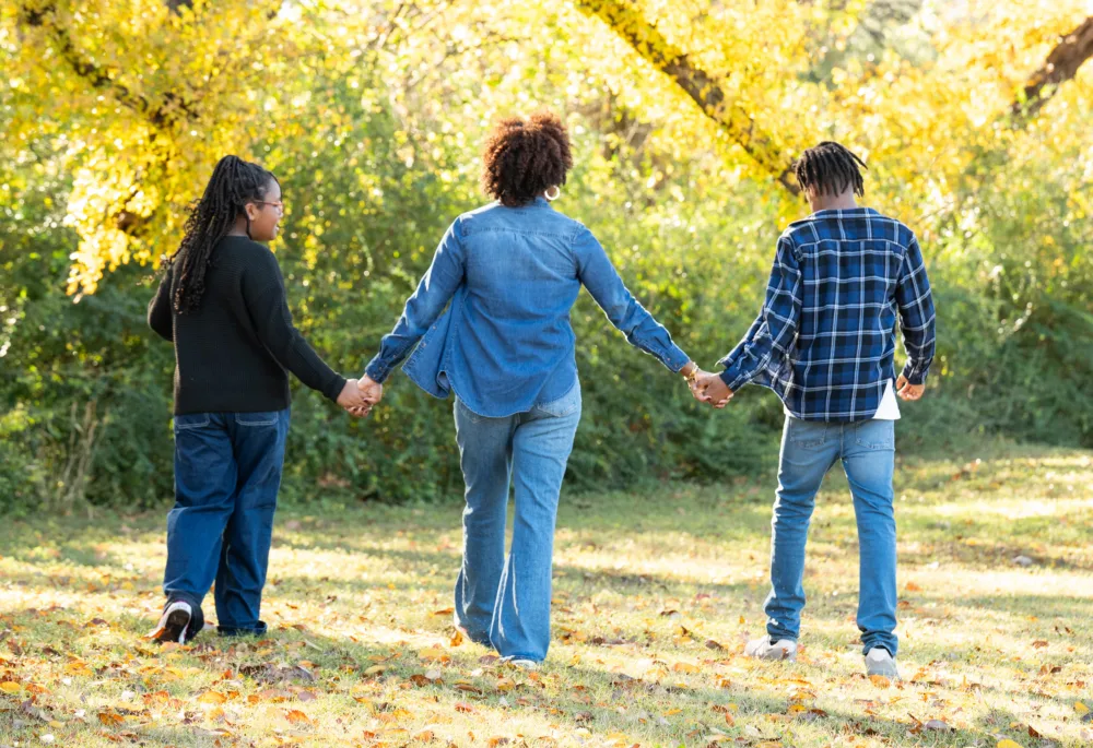 A family, seen from behind, including a woman and two teenagers, holding hands and walking together through a sunlit park with autumn foliage. The stylish and casual clothing they wear demonstrates the possibilities available through sustainable thrifting in South Carolina.