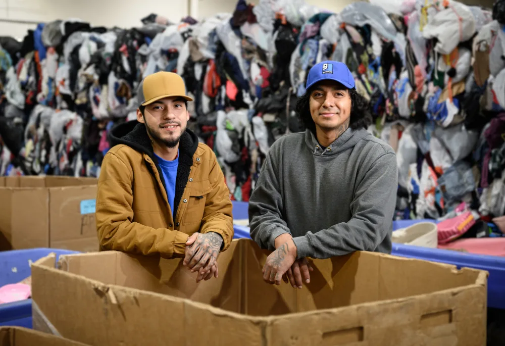 Two Palmetto Goodwill team members standing in front of large stacks of clothing donations, highlighting sustainable thrifting in South Carolina.