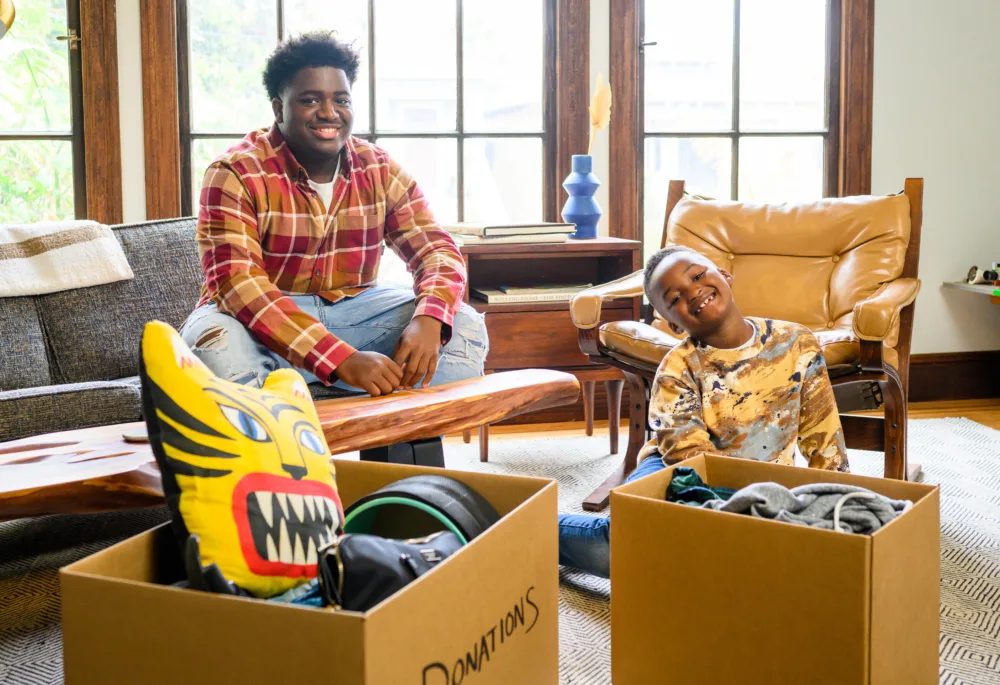A young man and a smiling child in a living room with boxes labeled for donation (containing a tiger-shaped pillow and home goods) to support sustainable thrifting in South Carolina.