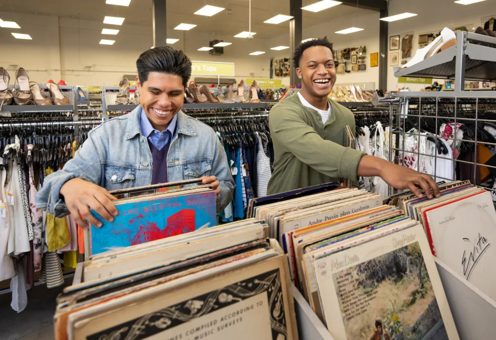 Two young men smiling and browsing through a curated selection of vintage vinyl records in a brightly lit, modern retail environment (promoting sustainable thrifting in South Carolina).