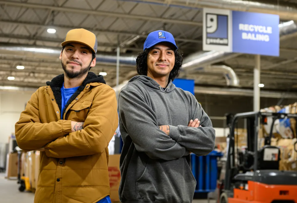 Two Palmetto Goodwill team members standing in front of a recycling baling station (representing the organization's commitment to sustainable thrifting in South Carolina).