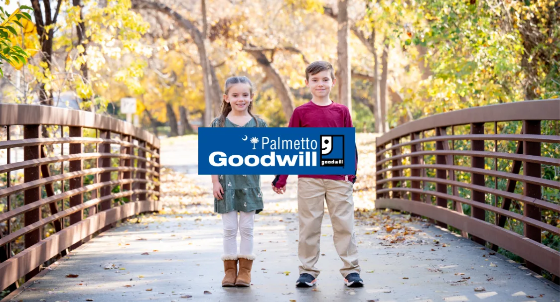 Two children stand hand-in-hand on an outdoor wooden bridge surrounded by autumn trees, with a blue Palmetto Goodwill logo banner overlaid across the center.