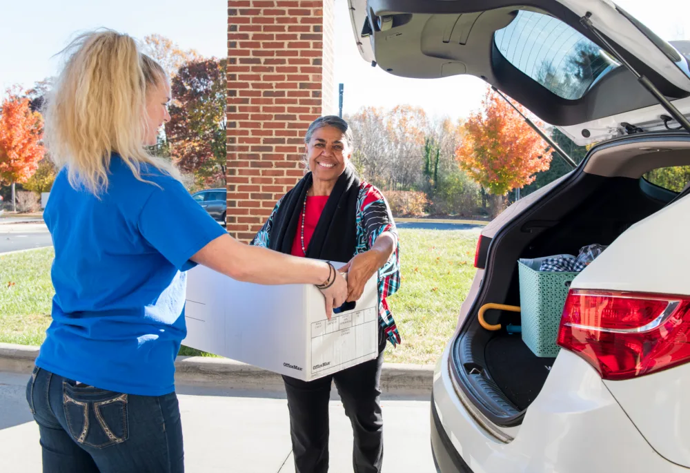 A Goodwill staff member in a blue shirt accepting a large white box of donations from a smiling woman next to her car.
