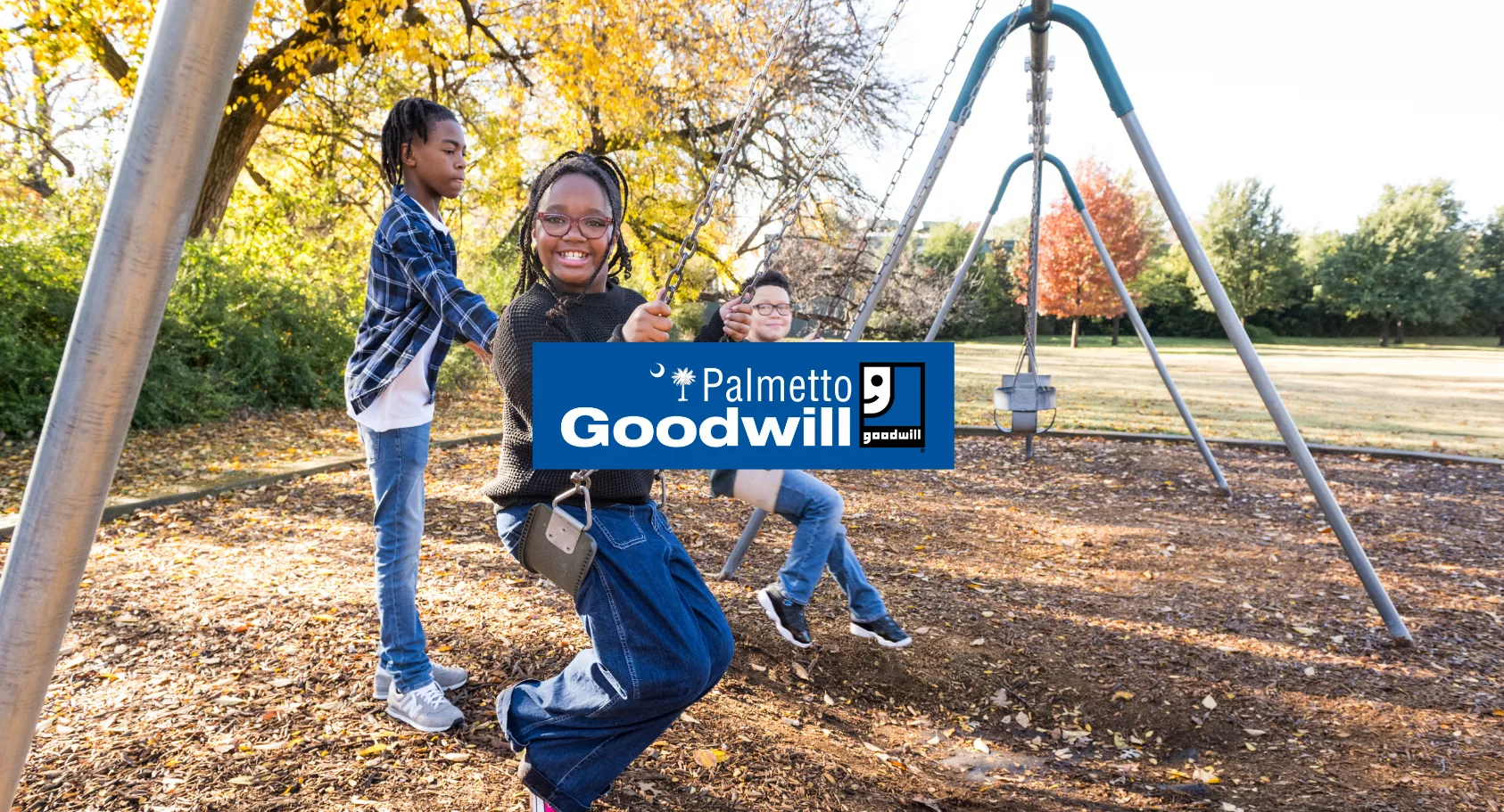 Three happy children playing on a swing set at a sunny park with the Palmetto Goodwill logo overlaid in the center.
