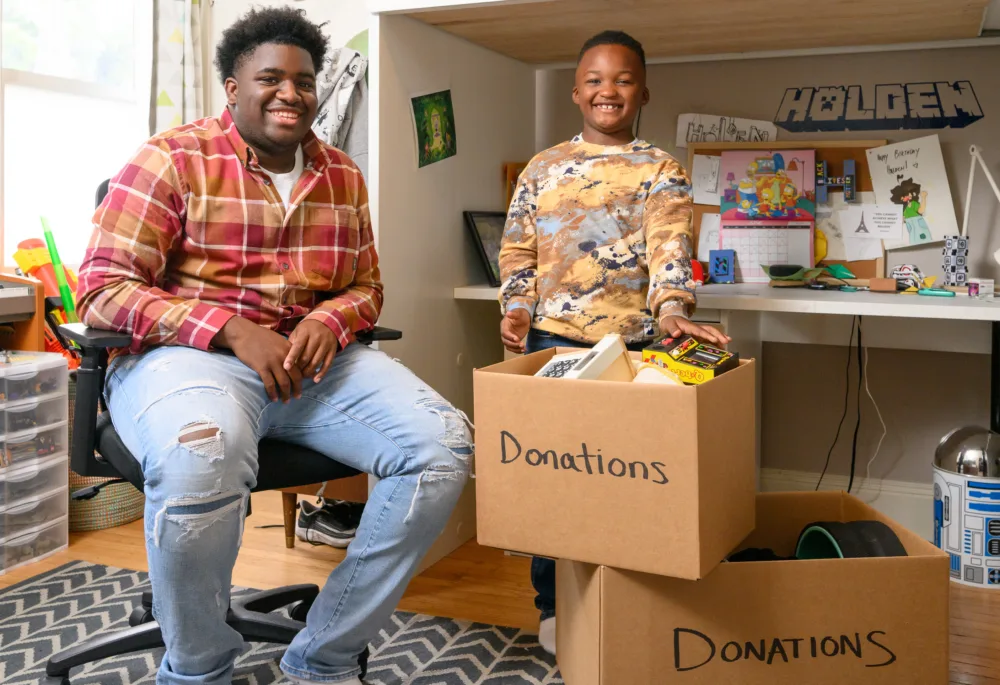 Two smiling young men in a bedroom posing next to cardboard boxes filled with toys and household items labeled "Donations."