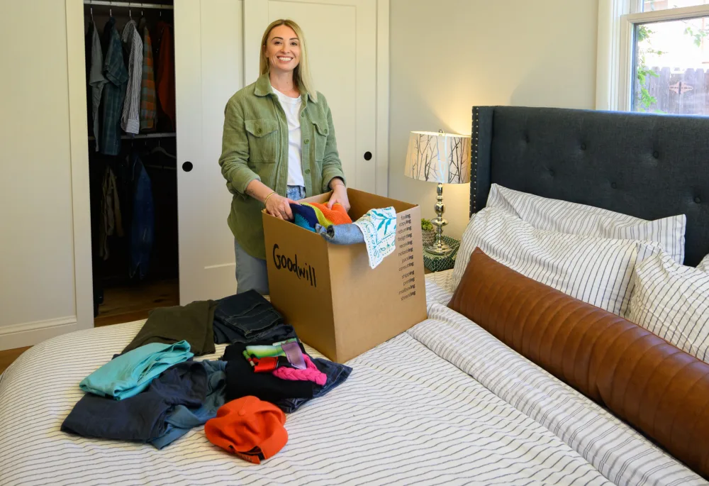 A smiling woman in a bedroom thoughtfully packs a cardboard box labeled "Goodwill" with colorful, gently used clothing and accessories, preparing them for donation to Palmetto Goodwill in South Carolina.