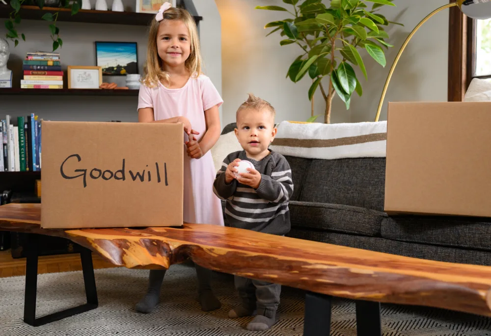 A young girl and a toddler boy standing in a living room next to a cardboard box labeled "Goodwill" on a wooden bench.